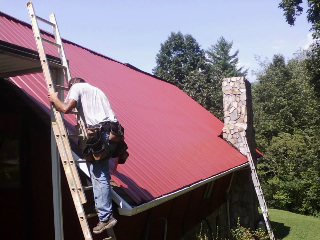 Roofer on a ladder beside a red metal roof and stone chimney — Soffit & Fascia Repair by Roofing Brothers in Old Lyme