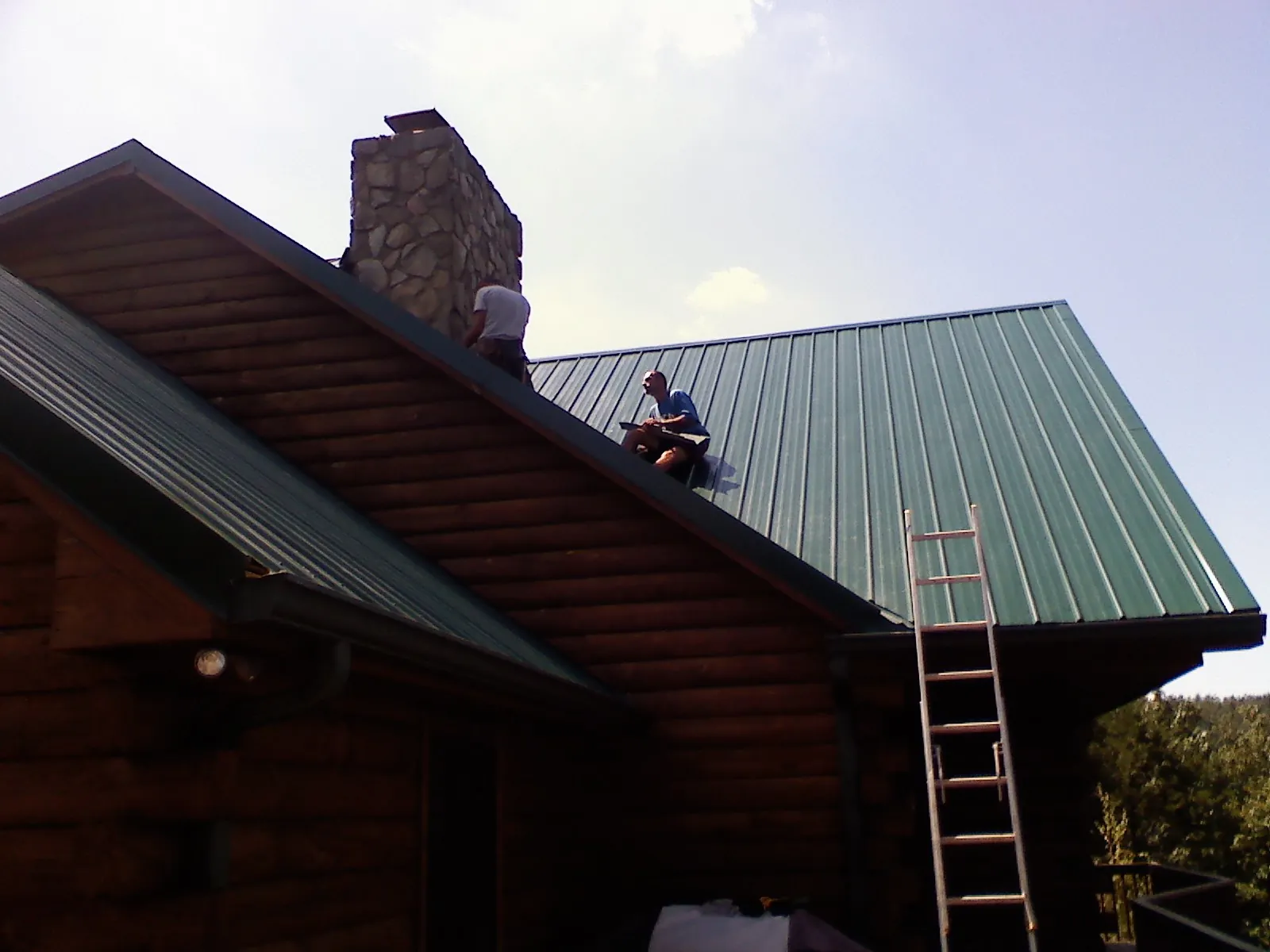 Roofers installing green metal panels beside a stone chimney on a log home in Old Lyme